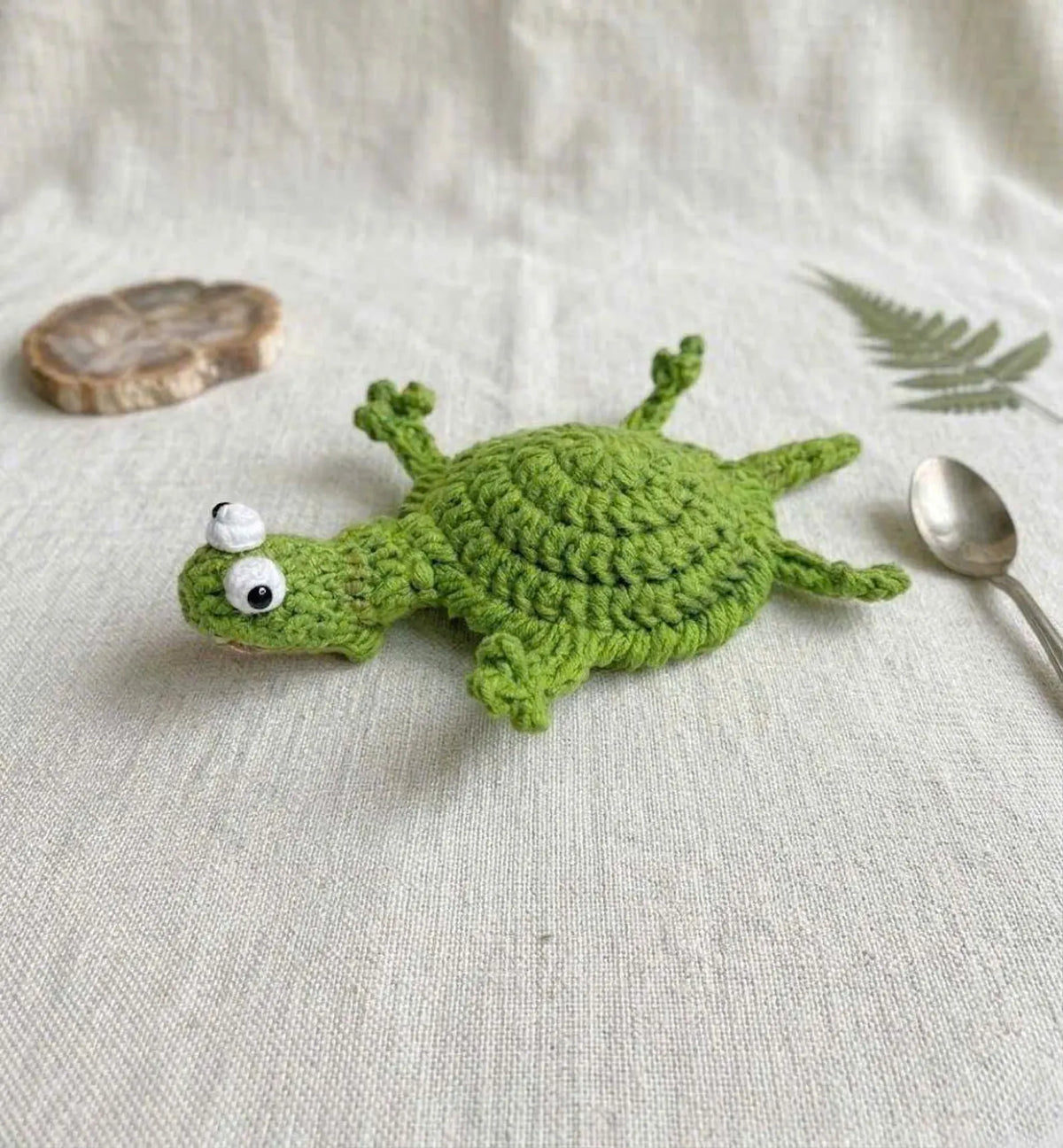 An aesthetic flat-lay table arrangement featuring the By1994 green handmade crochet lizard coaster laid flat on natural linen fabric beside a vintage dessert spoon, a dried botanical leaf, and a rustic wood coaster.