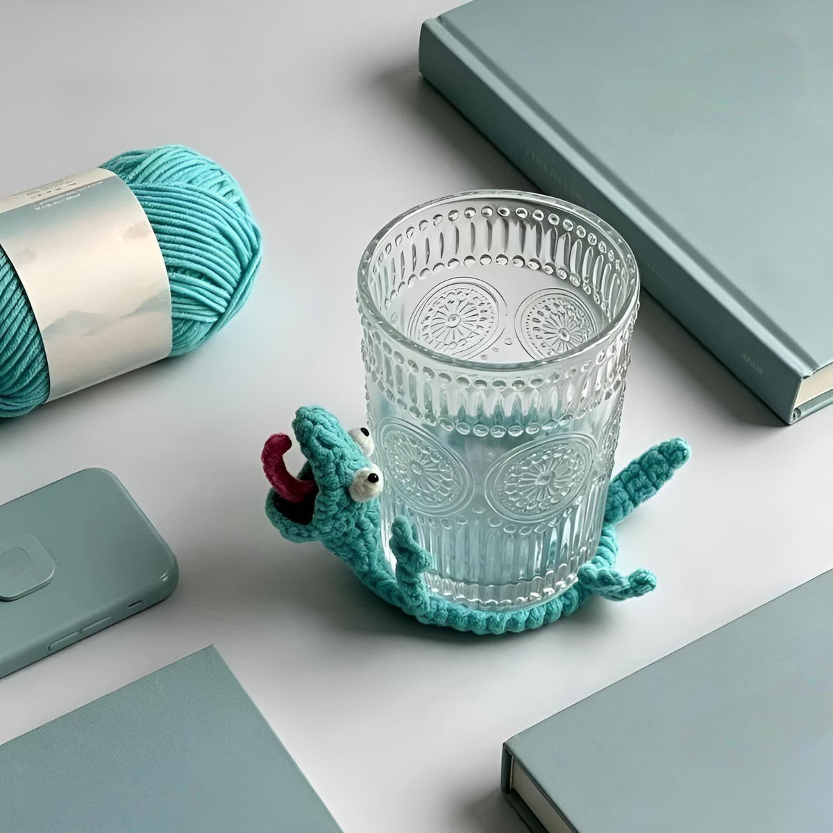 A modern workspace lifestyle setup showing an embossed clear glass tumbler resting on the By1994 blue crochet lizard coaster, positioned next to minimalist hardcover books and a cotton yarn skein on a desk.