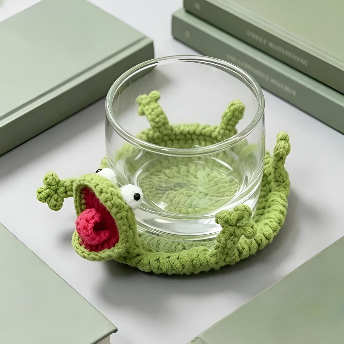A modern workspace lifestyle setup showing an embossed clear glass tumbler resting on the By1994 green crochet lizard coaster, positioned next to minimalist hardcover books and a cotton yarn skein on a desk.