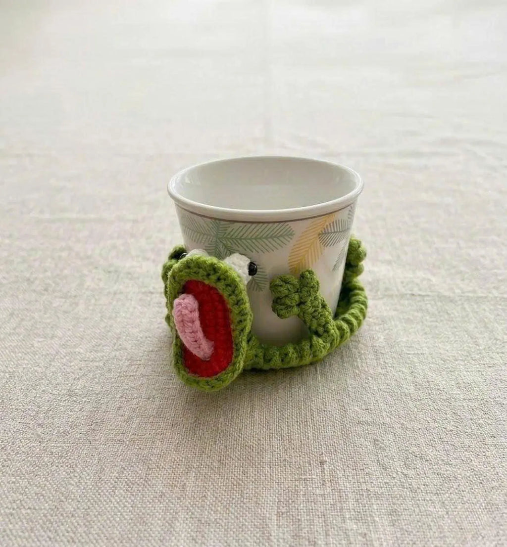 A close-up lifestyle shot of the By1994 sage green crochet lizard coaster gently wrapping around the base of a white patterned ceramic coffee mug on a neutral textured linen tablecloth.