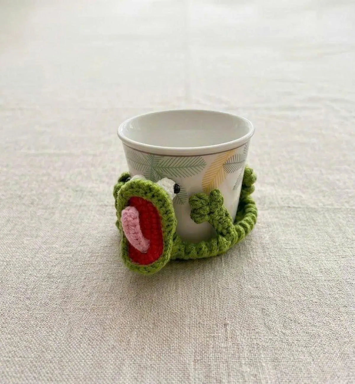A close-up lifestyle shot of the By1994 sage green crochet lizard coaster gently wrapping around the base of a white patterned ceramic coffee mug on a neutral textured linen tablecloth.