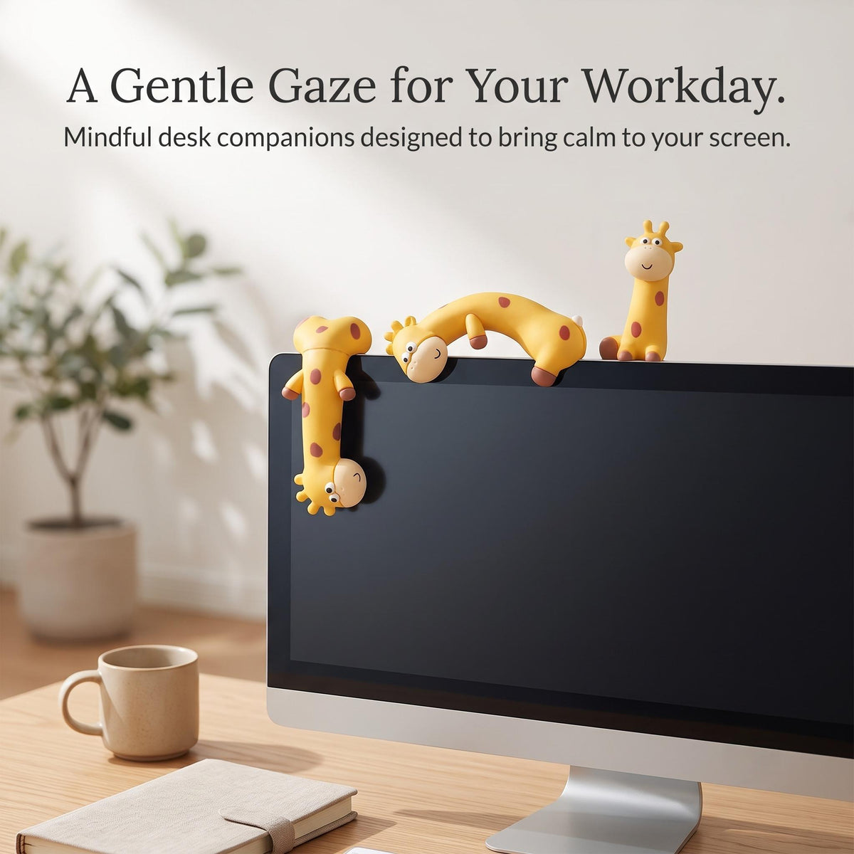 A serene workspace lifestyle setup showing three By1994 mini giraffe figurines resting along the top rim of a computer monitor beside a coffee mug and notebook, with text overlay reading A Gentle Gaze for Your Workday. Mindful desk companions designed to bring calm to your screen.